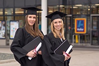 Zwei Masterabsolventinnen in klassischer Kluft mit Haube und Abschlussdokumenten in der Hand lächeln in die Kamera.